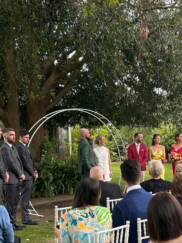 Wedding ceremony in a garden with bride and groom under a white arch and grooms men and bridesmaids either side. Wedding party seated in front looking on.