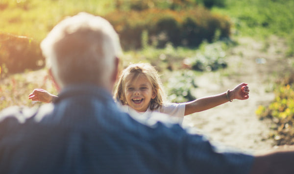 A young granddaughter running excitedly towards her grandfather with open arms, both smiling warmly as they prepare to embrace, set against a bright outdoor background.
