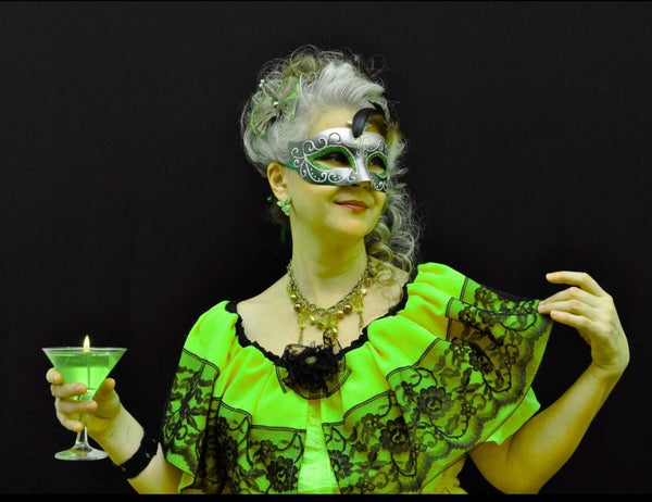 Person wearing a green dress with black lace details and a mask, holding a green cocktail.