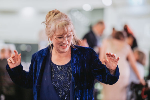 A woman wearing a blue jacket and glasses dancing joyfully, her arms raised in motion, with a bright and lively expression on her face.